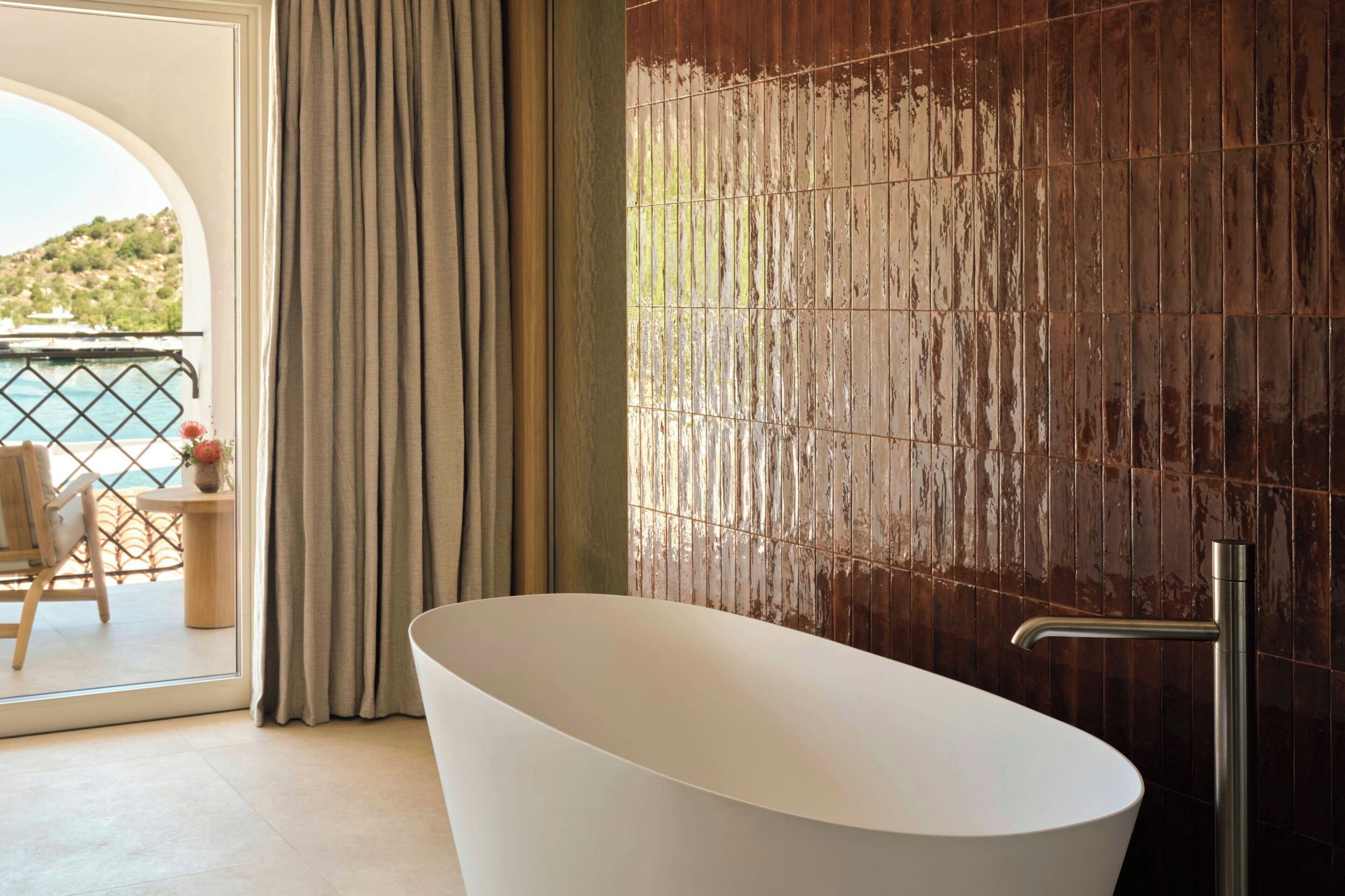 A modern bathroom with a freestanding bathtub, a view of the sea through a glass door, and a balcony with outdoor furniture.