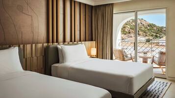 Habitación de hotel con dos camas, un ventanal con cortinas, vista a un paisaje montañoso y un balcón con sillas.