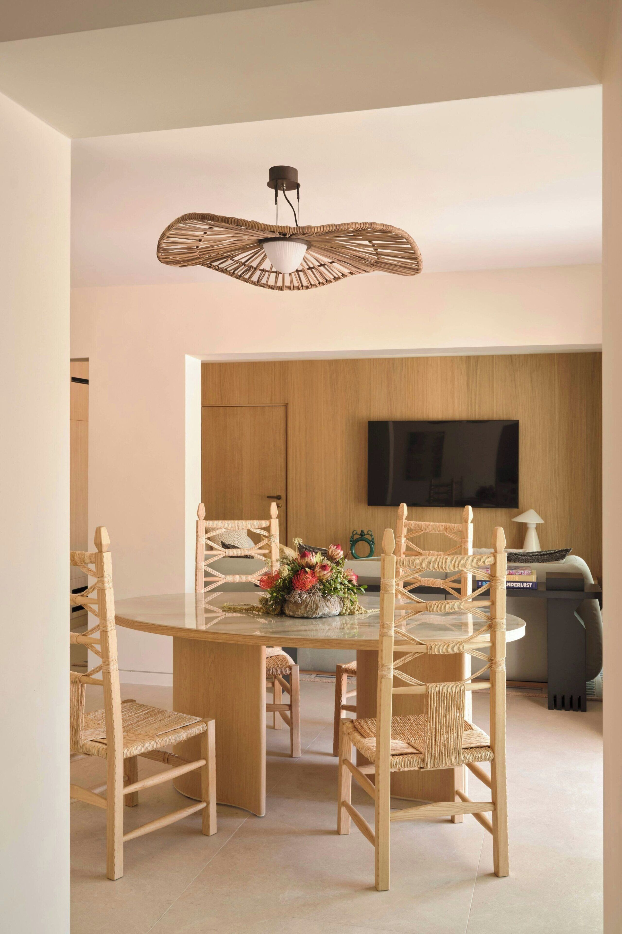 Un coin repas avec une table ronde, quatre chaises en osier, un luminaire central et un téléviseur en arrière-plan.