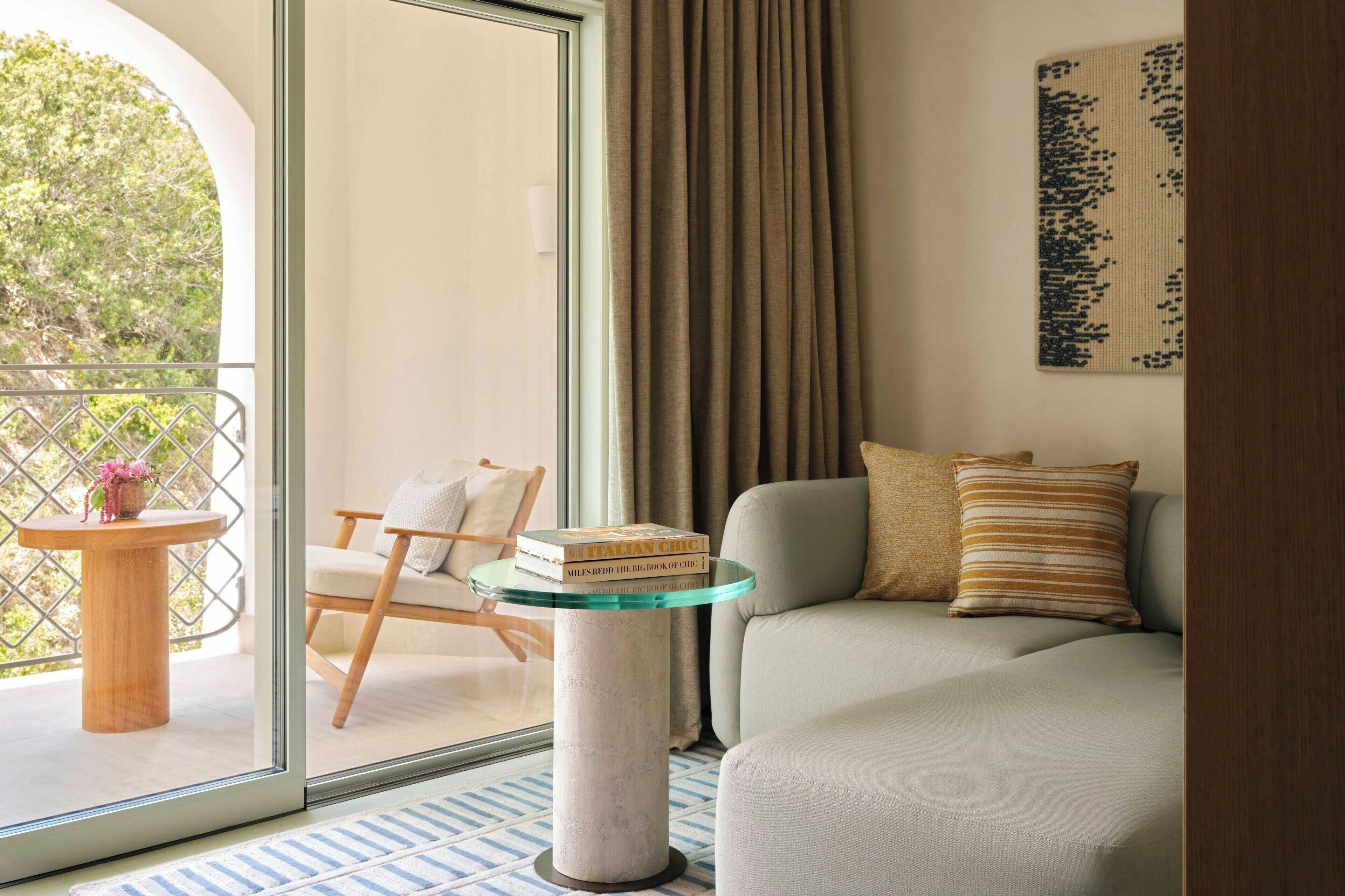 Una habitación con una mesa de vidrio, un sofá, una silla, un ventanal con cortinas y vistas a zonas verdes.