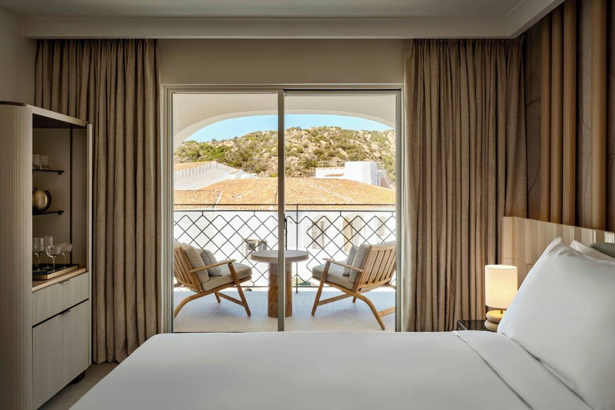 A hotel room with a large bed, a desk with a lamp, a chair, and a balcony with a table and chairs.
