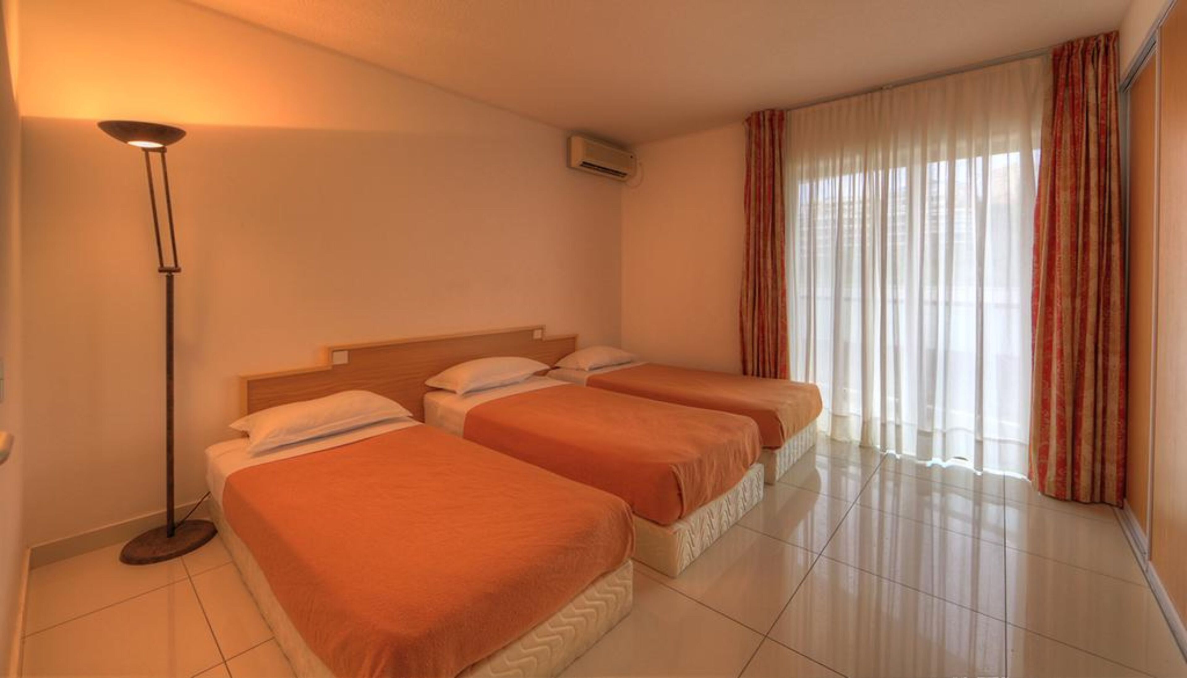Habitación de hotel con tres camas individuales, una lámpara de pie, una ventana con cortinas e un equipo de aire acondicionado.