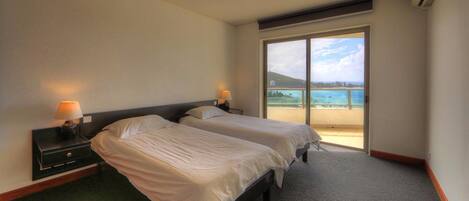 A hotel room with two beds, a nightstand with a lamp, and a large sliding glass door leading to a balcony with a view of the sea.