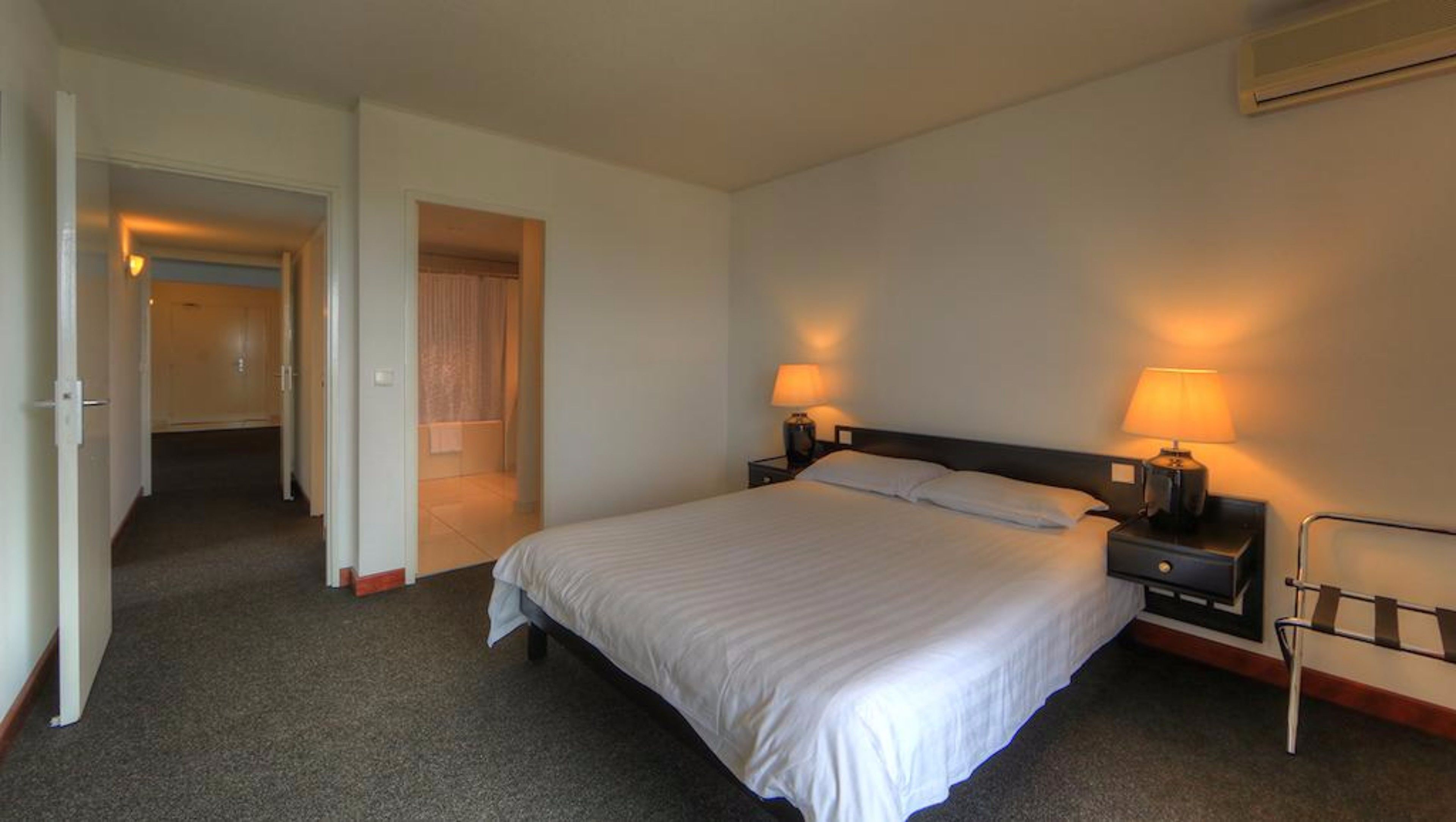 A hotel room with a large bed, two bedside tables with lamps, a small desk, and a chair. There is a door leading to a bathroom and a hallway.