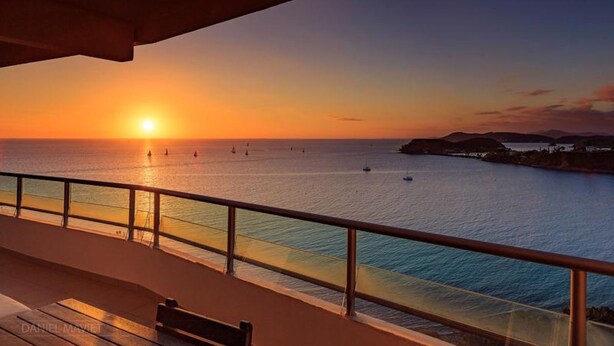 A sunset view from a balcony overlooking a calm sea with boats, a clear sky, and distant hills.