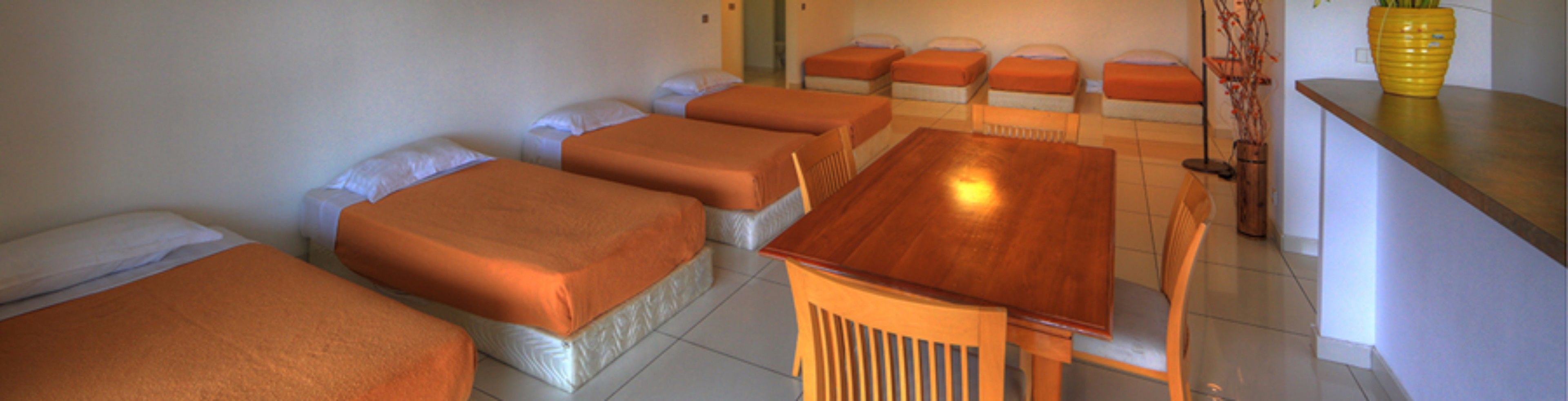 Una habitación con varias camas dispuestas en fila, una mesa de madera y un jarrón amarillo con ramas.