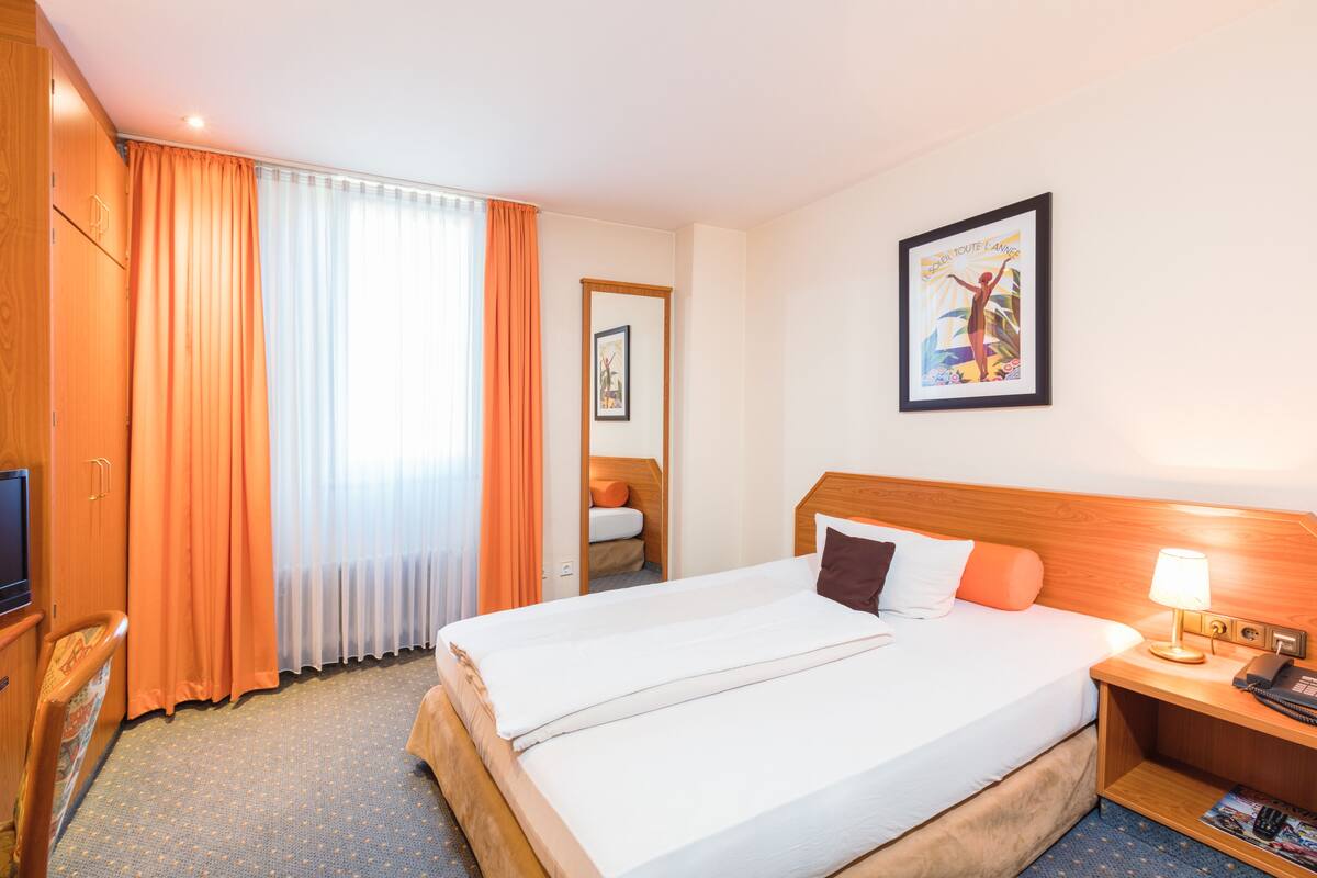 A hotel room with a double bed, a wooden headboard, a bedside table with a lamp, a TV, and a window with orange curtains.
