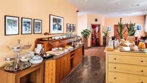 Espaço de café da manhã de hotel, com buffet, quadros nas paredes e um relógio.
