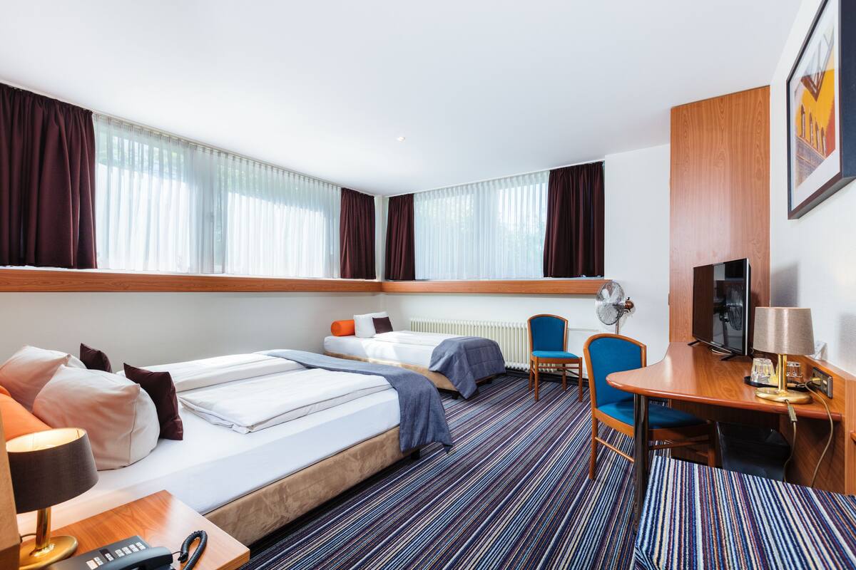 A hotel room with two beds, a desk, a chair, a TV, and a window with curtains.
