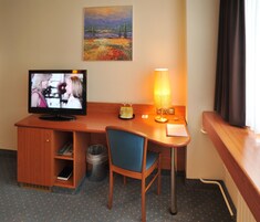 A hotel room with a wooden desk, a television, a lamp, a chair, and a painting on the wall.