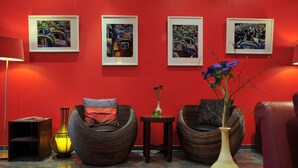 Um quarto de paredes vermelhas com três obras de arte emolduradas, duas cadeiras de vime, uma pequena mesa com um vaso e um abajur.