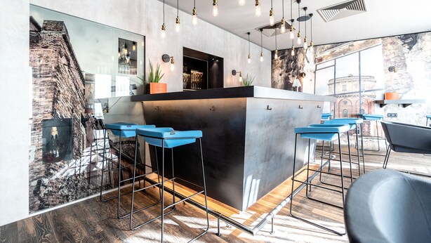 Zona de bar moderna con mostrador central, taburetes azules, lámparas colgantes y un amplio ventanal con vistas a un edificio histórico.