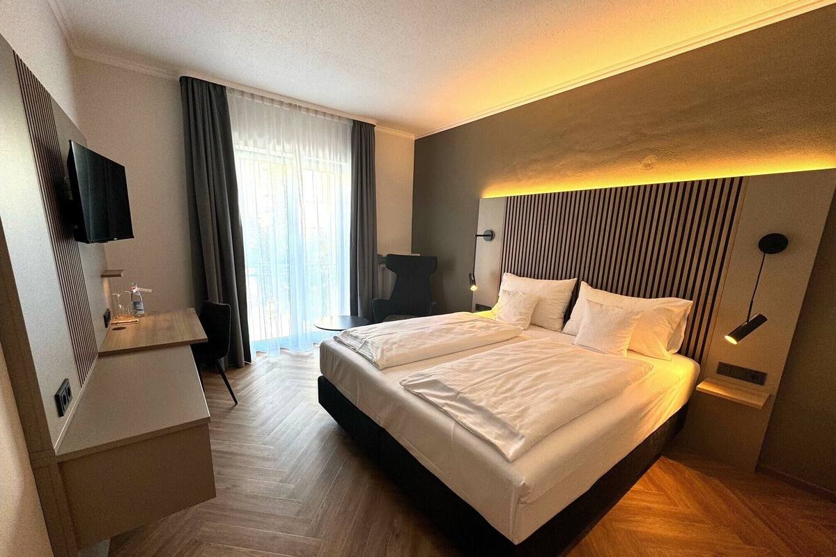 A hotel room with a large bed, a desk, a chair, a TV, and a window with curtains.