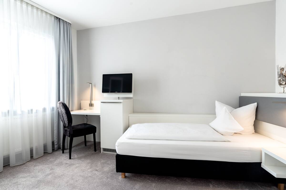A hotel room with a bed, a desk, a chair, a TV, and a window with curtains.