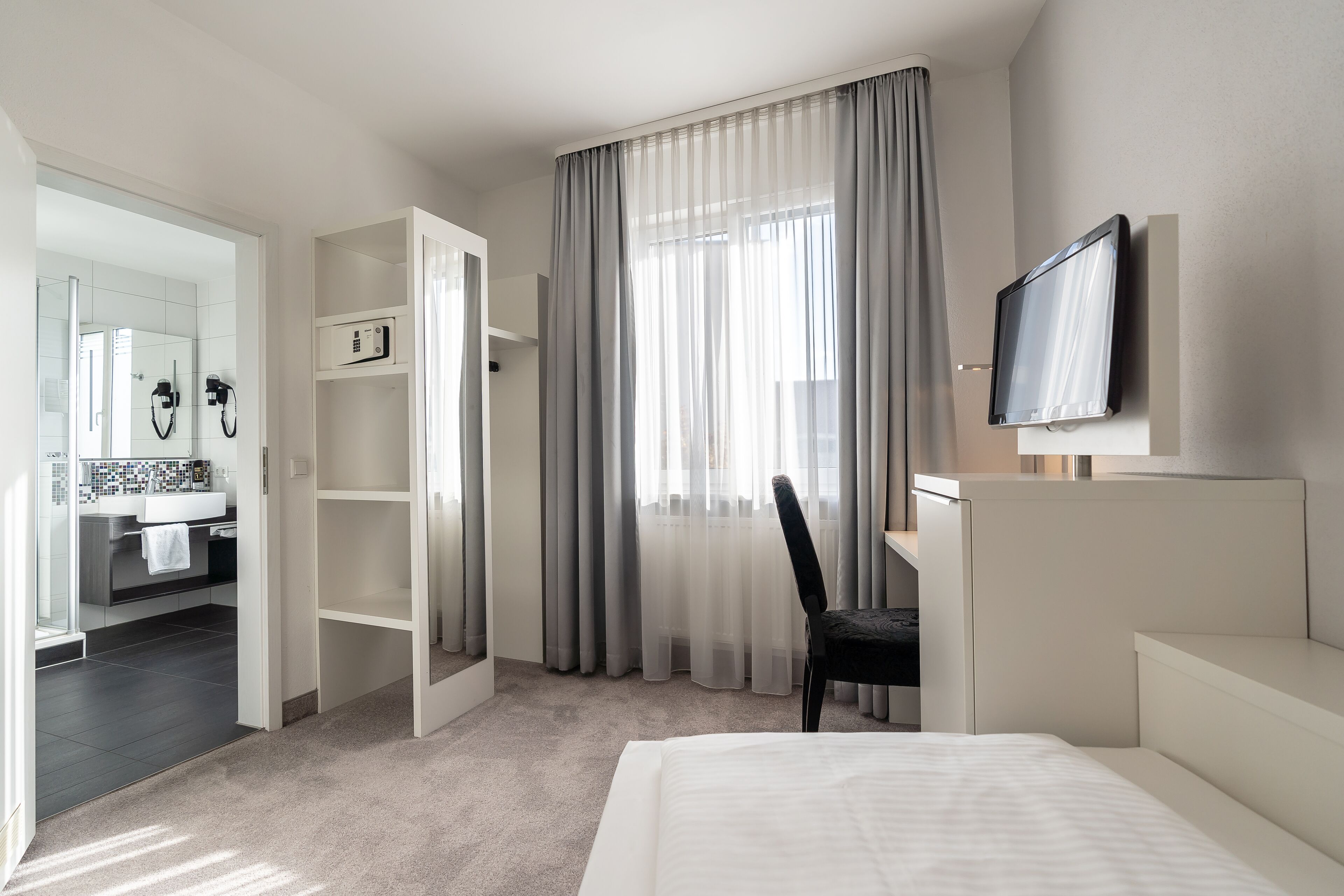 Una habitació d'hotel amb un llit, una taula de treball, una cadira, una televisió, un armari i un bany visible a través d'una porta oberta.