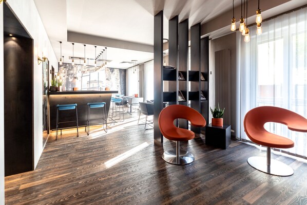 A modern interior with a wooden floor, a curved orange chair, a bar area, and a large window with natural light. - Altstadthotel (Ingolstadt)