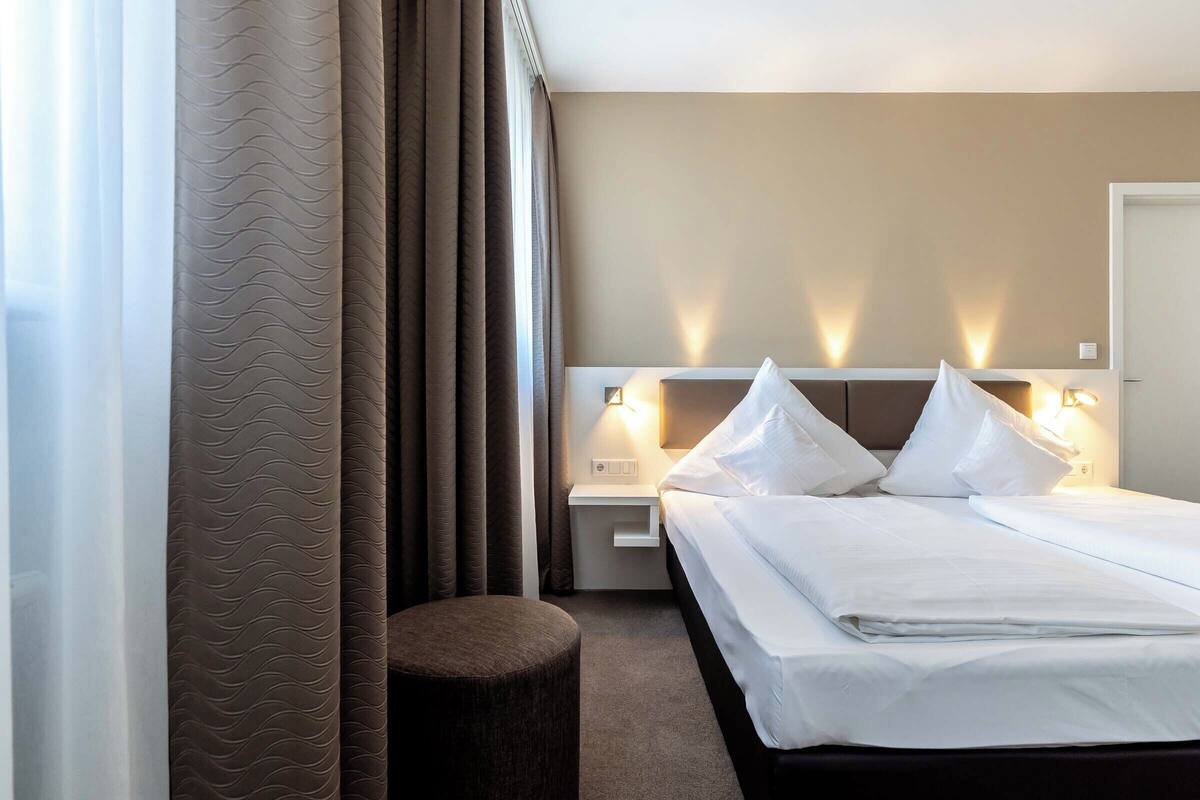 A hotel room with a neatly made bed, a bedside table with a lamp, and a view of the outside through curtains.