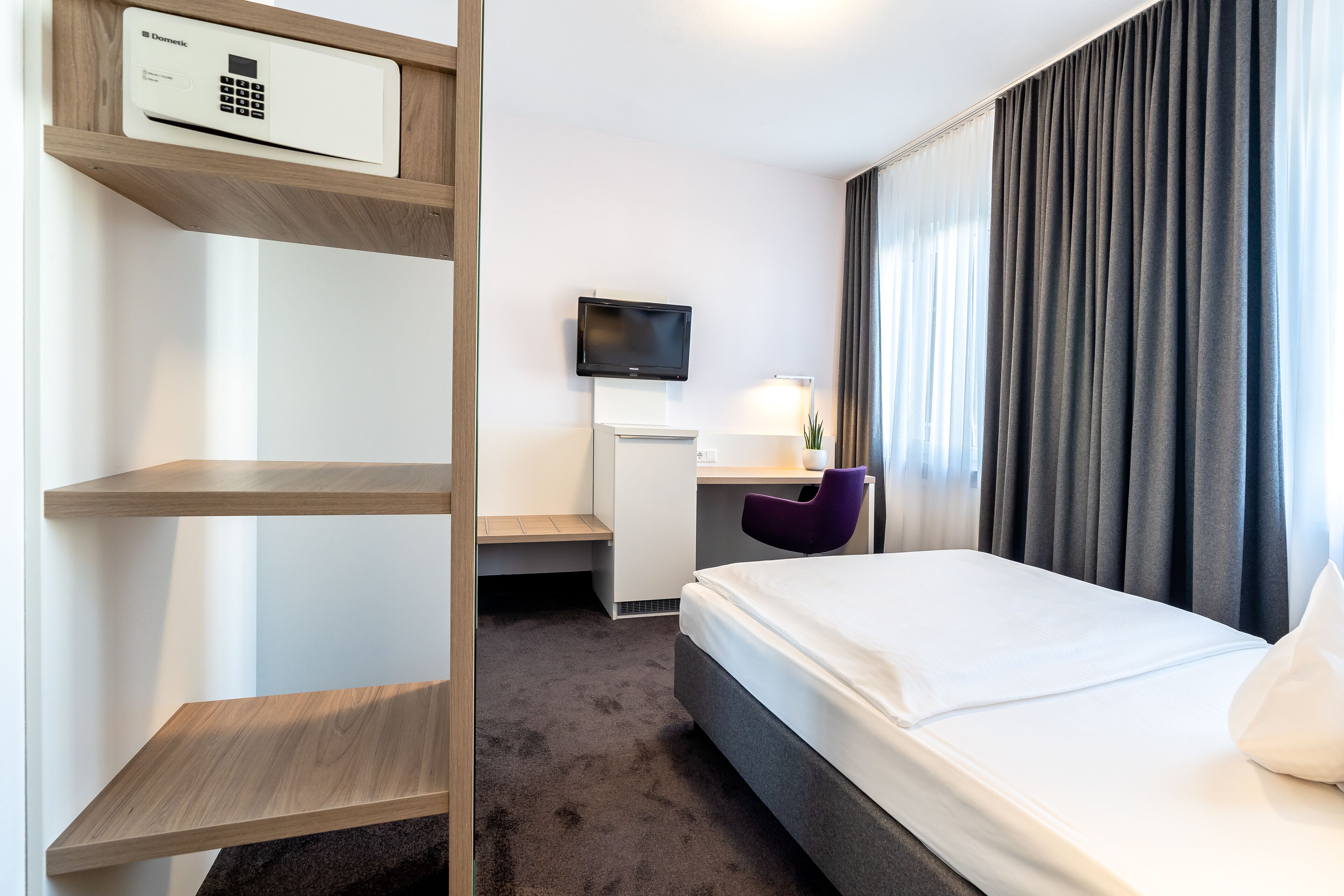 A hotel room with a bed, a desk with a computer, a TV, and a control panel on the wall.