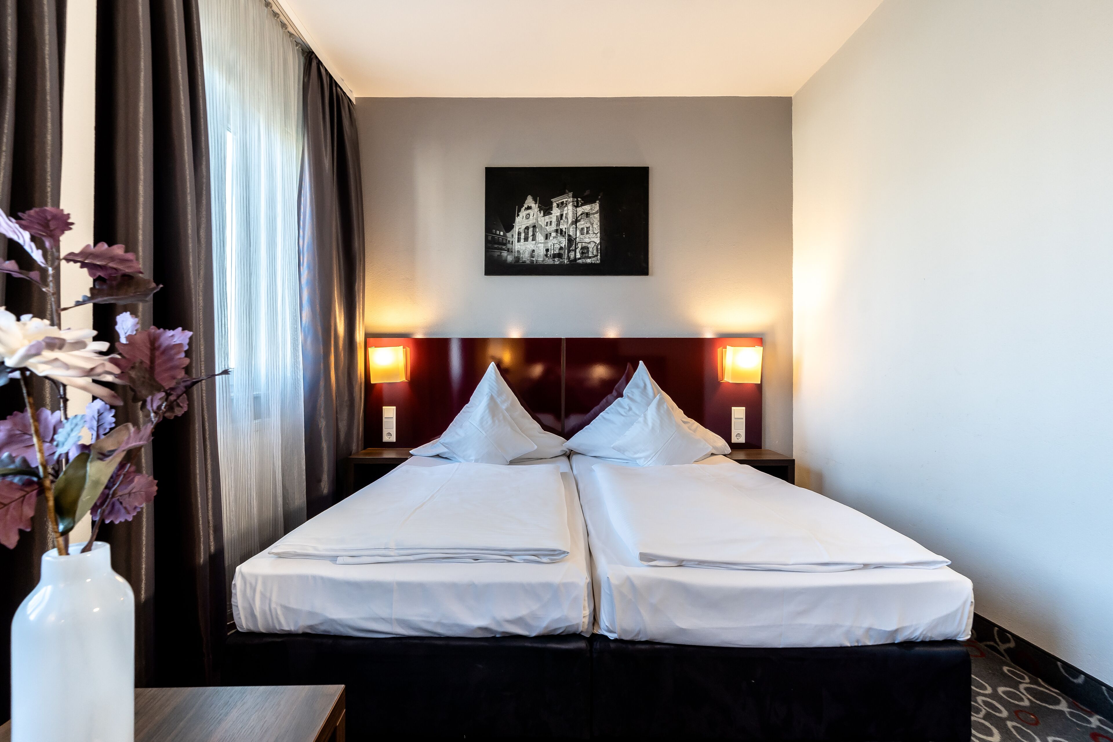 A hotel room with a double bed, two bedside tables, a vase with flowers, a painting on the wall, and a view of the cityscape.