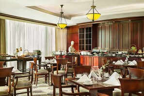 A hotel dining area with wooden furniture, a buffet setup, and a chef preparing food. - Metropolitan Palace (Sin El Fil)