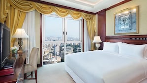 A hotel room with a large bed, two bedside lamps, a desk with a lamp, a chair, and a city view through the window.