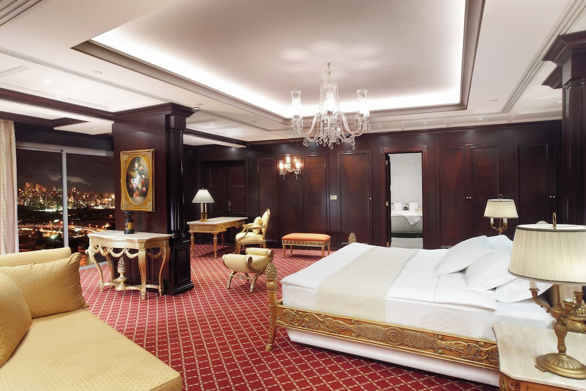 A luxurious hotel room with a large bed, a sitting area with a sofa and armchairs, a dining table, and a view of the cityscape.