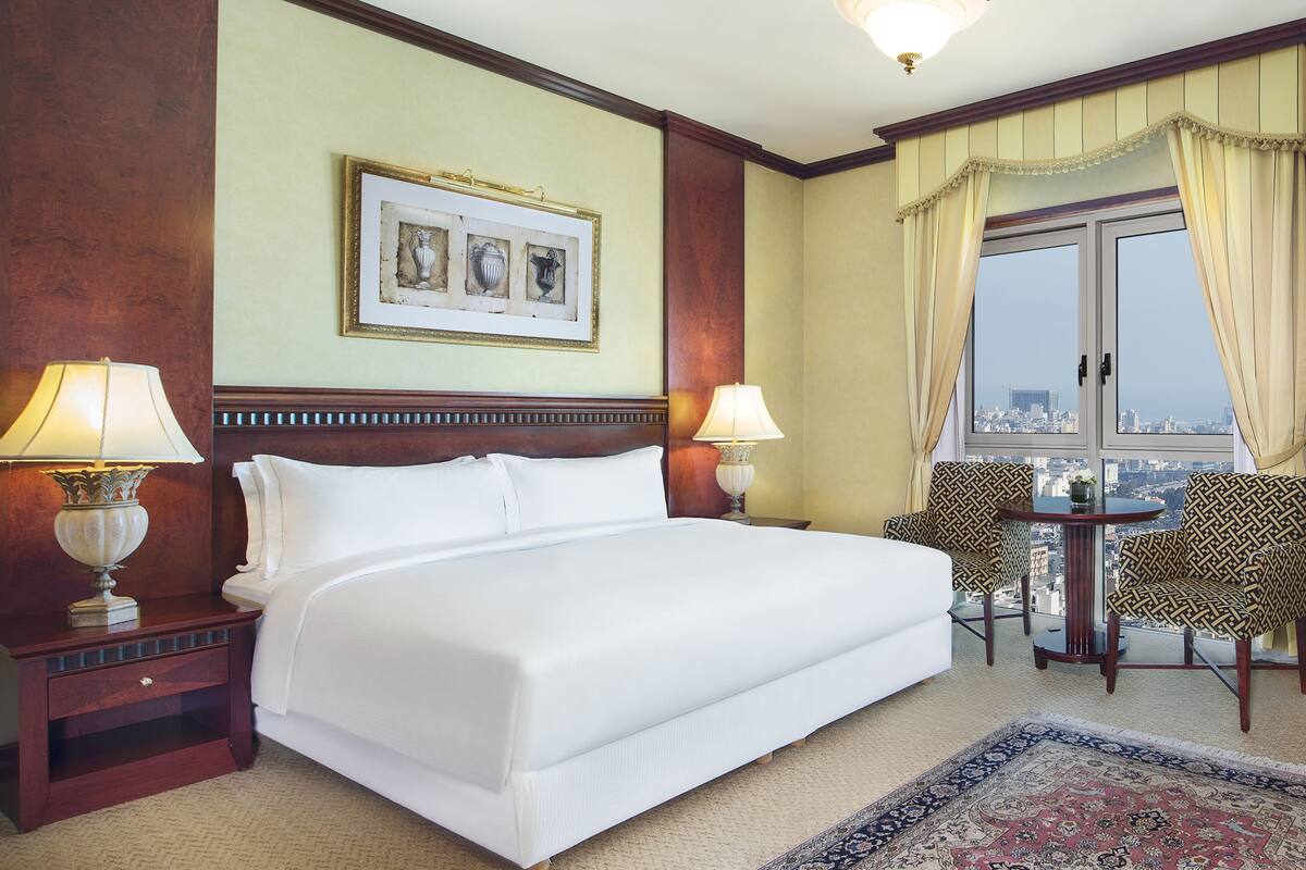 A hotel room with a large bed, two bedside lamps, a framed artwork above the bed, a view of the city through a window, and a small table with chairs.