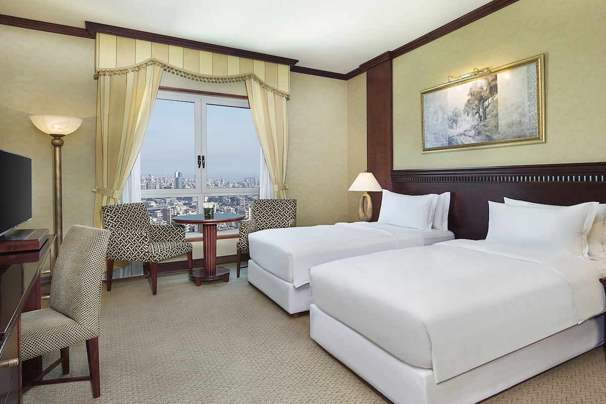 A hotel room with two beds, a desk with a lamp, a chair, and a large window with curtains.