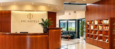 A hotel reception area with a wooden reception desk, a seating area with a sofa and armchairs, and a bookshelf with various items.