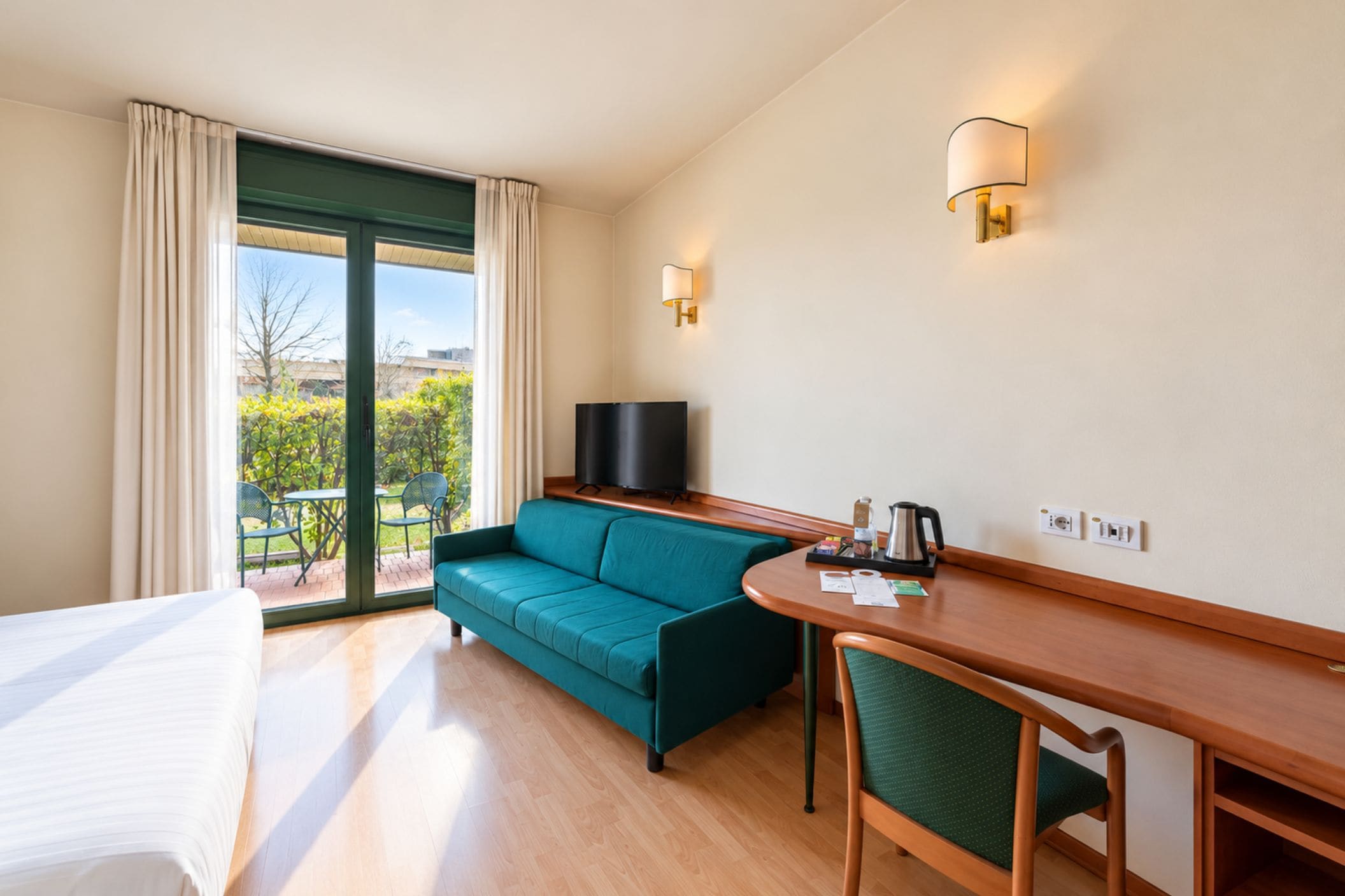 Una camera d'hotel con un letto, un divano color petrolio, una scrivania in legno, una televisione e vista sul verde.