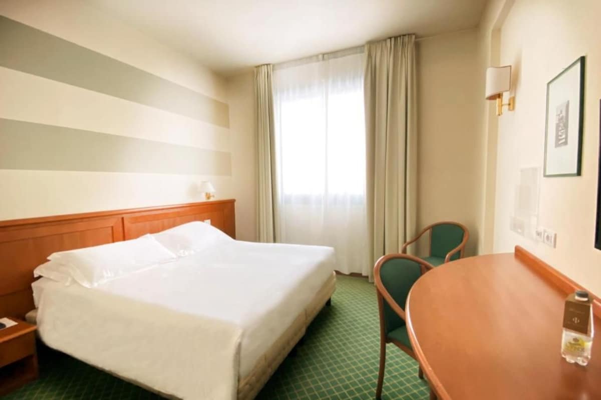 Ein Hotelzimmer mit einem großen Bett, einem Holfnachtkästchen, einem grün karierten Boden, einem runden Holztisch und einem grünen Stuhl.