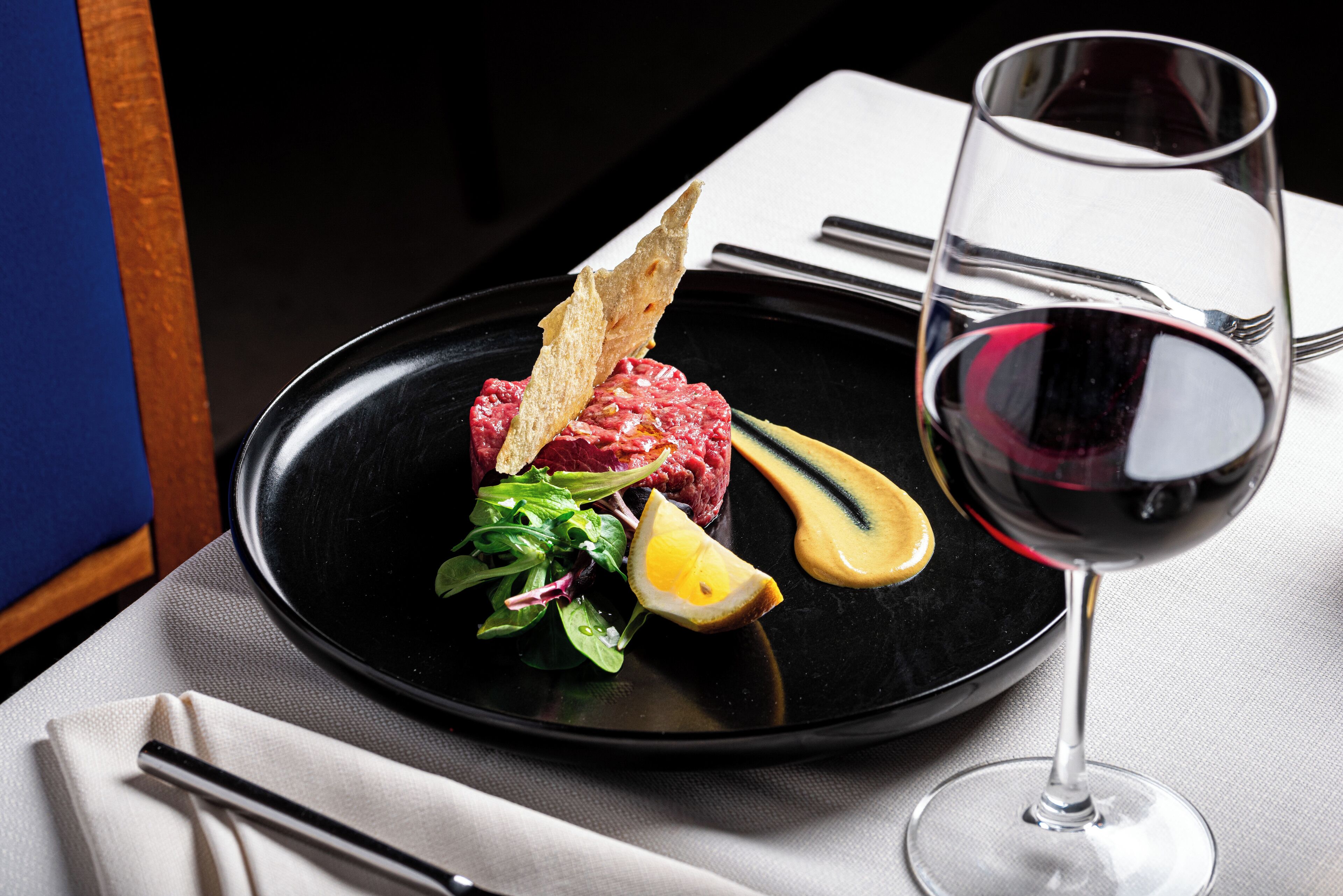 Un plato negro con un filete, limón y vegetales verdes, acompañado de una copa de vino tinto y un tenedor sobre una servilleta blanca.
