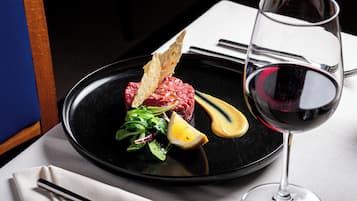 Un plato negro con un filete, limón y vegetales verdes, acompañado de una copa de vino tinto y un tenedor sobre una servilleta blanca.