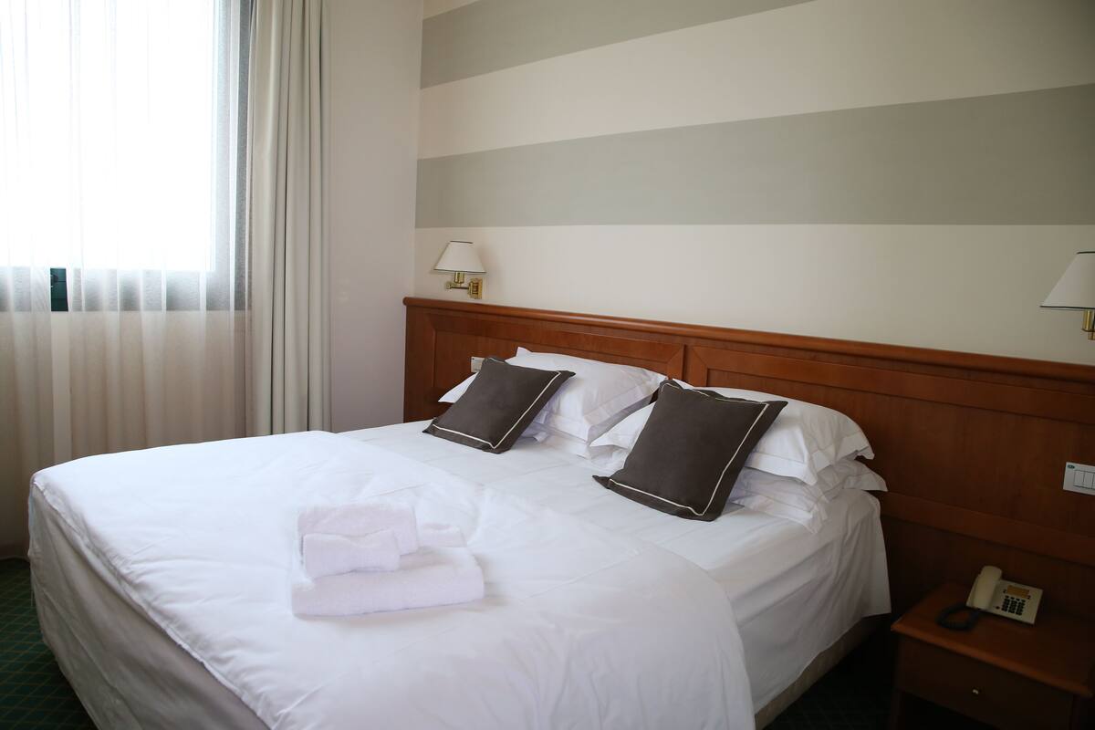 Ein Hotelzimmer mit einem großen Bett, weißer Bettwäsche, zwei dunklen Kissen, einer Nachttischlampe und einem Telefon auf dem Nachttisch.