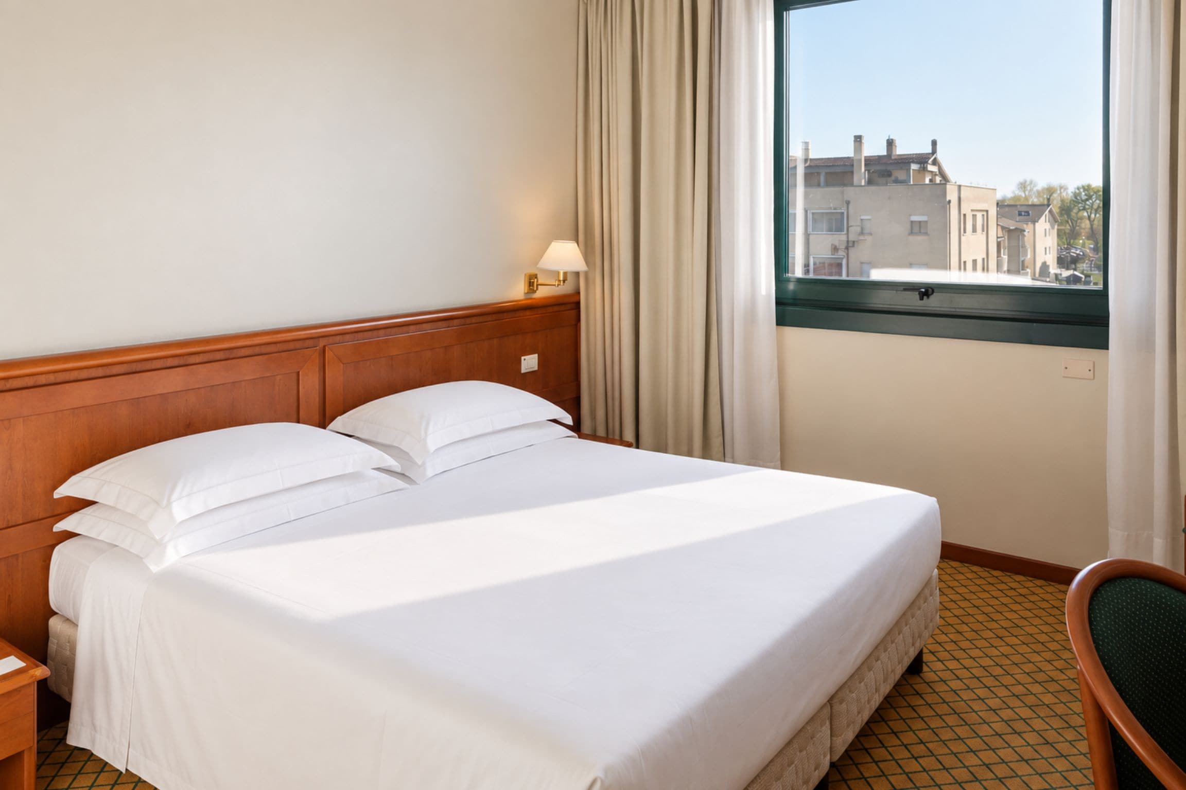 Habitación de hotel con cama, mesita de noche, una silla, ventana con cortinas y vista a edificios.