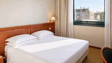 Habitación de hotel con cama, mesita de noche, una silla, ventana con cortinas y vista a edificios.