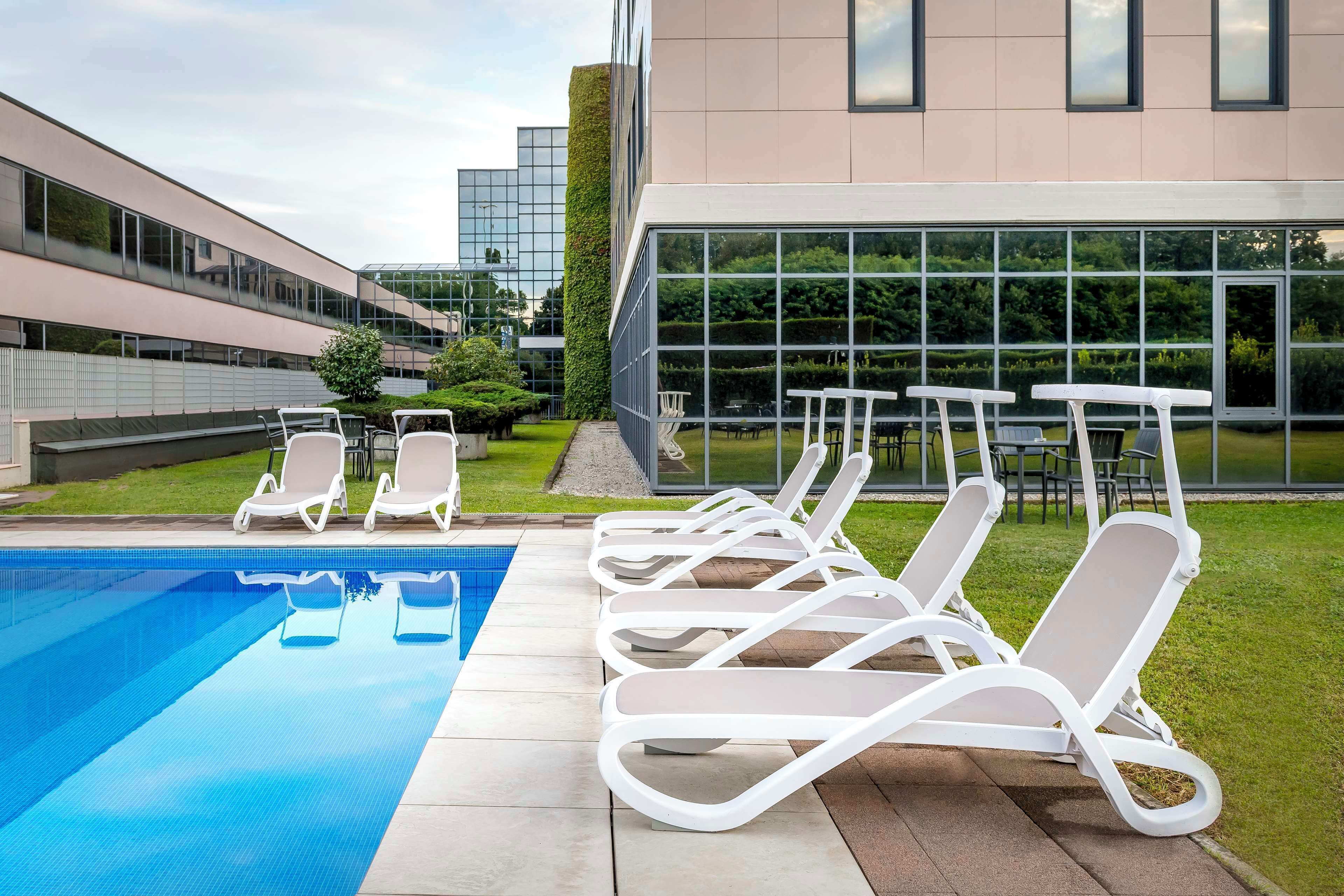 A swimming pool with lounge chairs and tables, surrounded by a modern building with large windows.