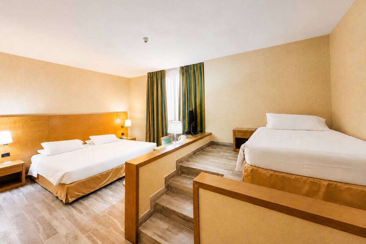 A hotel room with two beds, a wooden headboard, a desk with a chair, a TV, and a window with curtains.