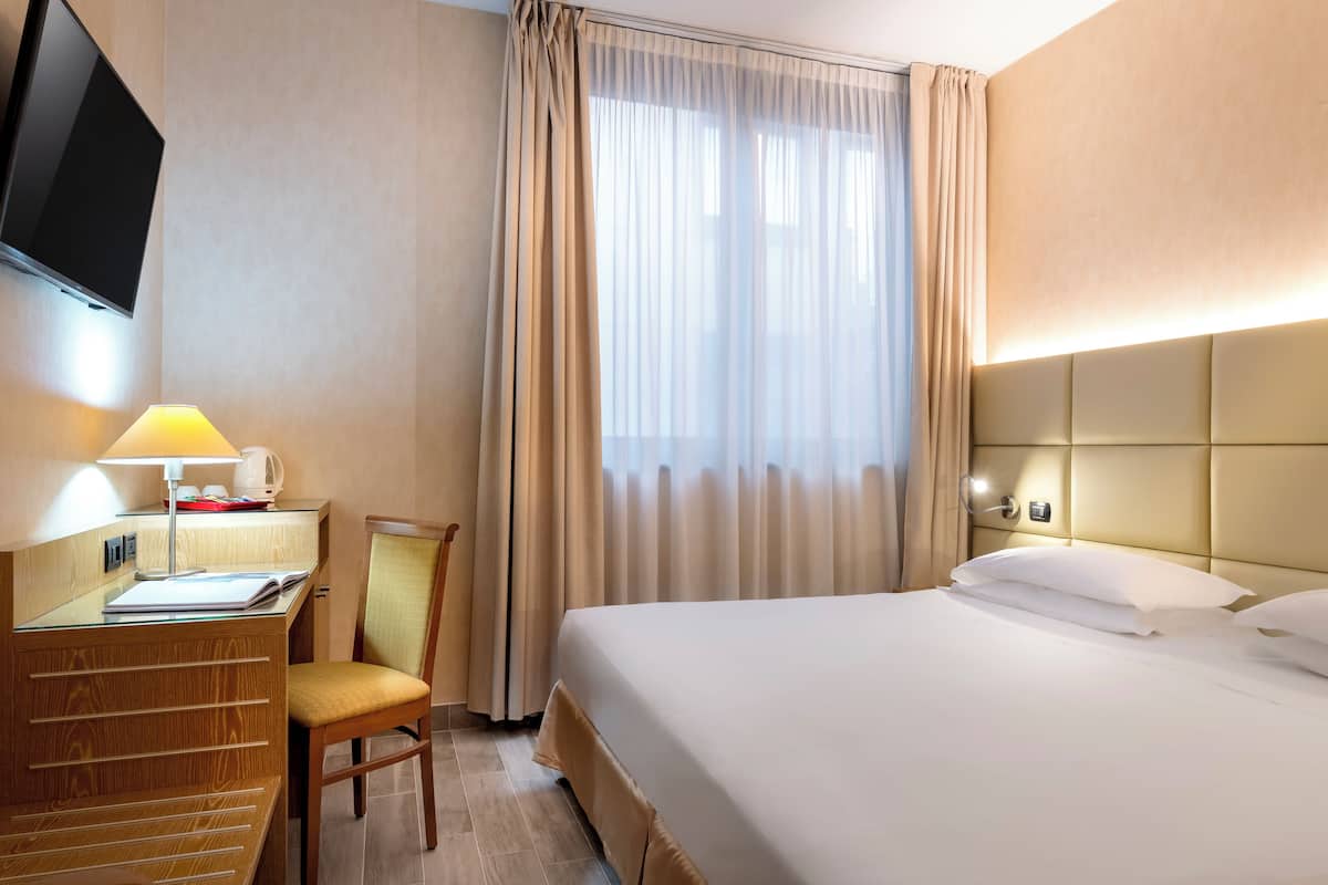 A hotel room with a bed, a desk with a lamp, a chair, and a TV mounted on the wall.