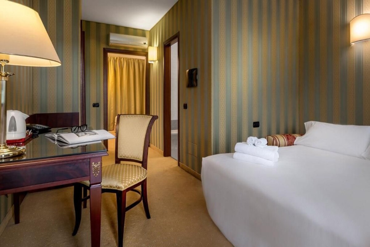 A hotel room with a bed, a desk with a lamp, a chair, and a door leading to another room.