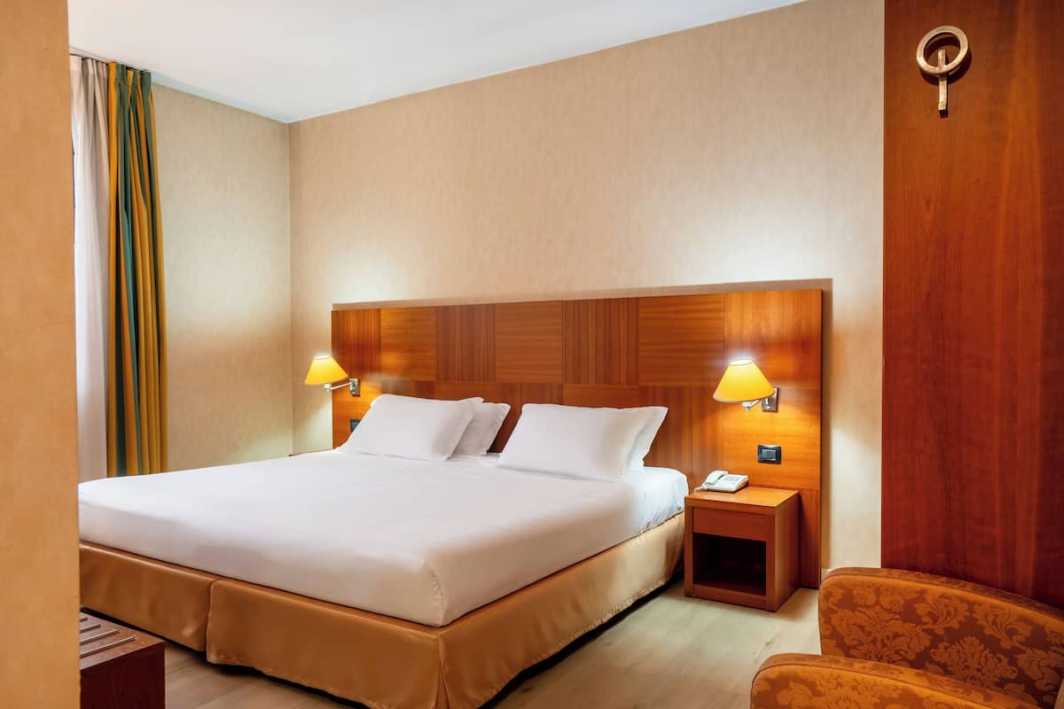 A hotel room with a large bed, two bedside lamps, a wooden headboard, a nightstand, and a patterned armchair.