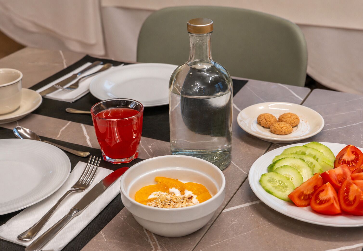 Una mesa dispuesta con platos, cubiertos, un vaso de bebida roja, una botella de agua, un bol con rodajas de naranja y granola, y un plato con rodajas de pepino y tomate.