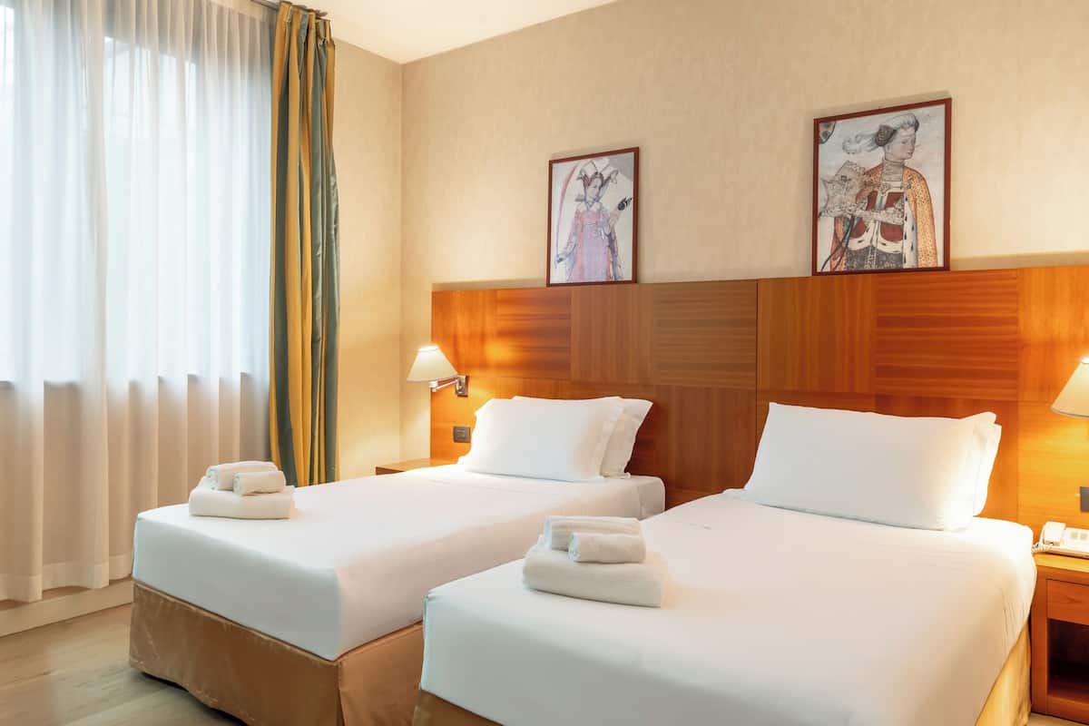 A hotel room with two beds, a wooden headboard, a bedside table with a lamp, and artwork on the wall.