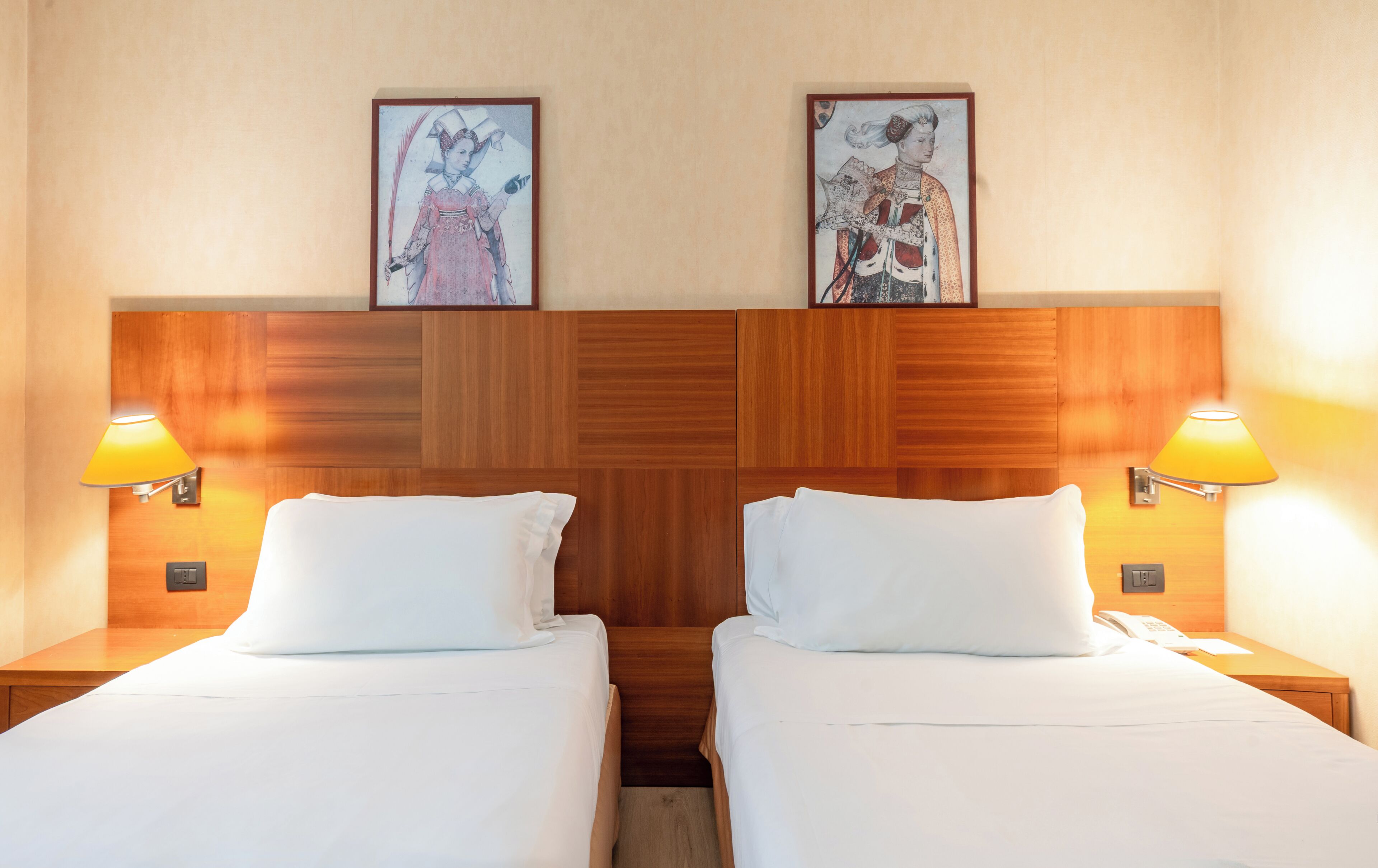 Habitación de hotel con dos camas, cabecero de madera, dos lámparas de noche y dos cuadros en la pared.