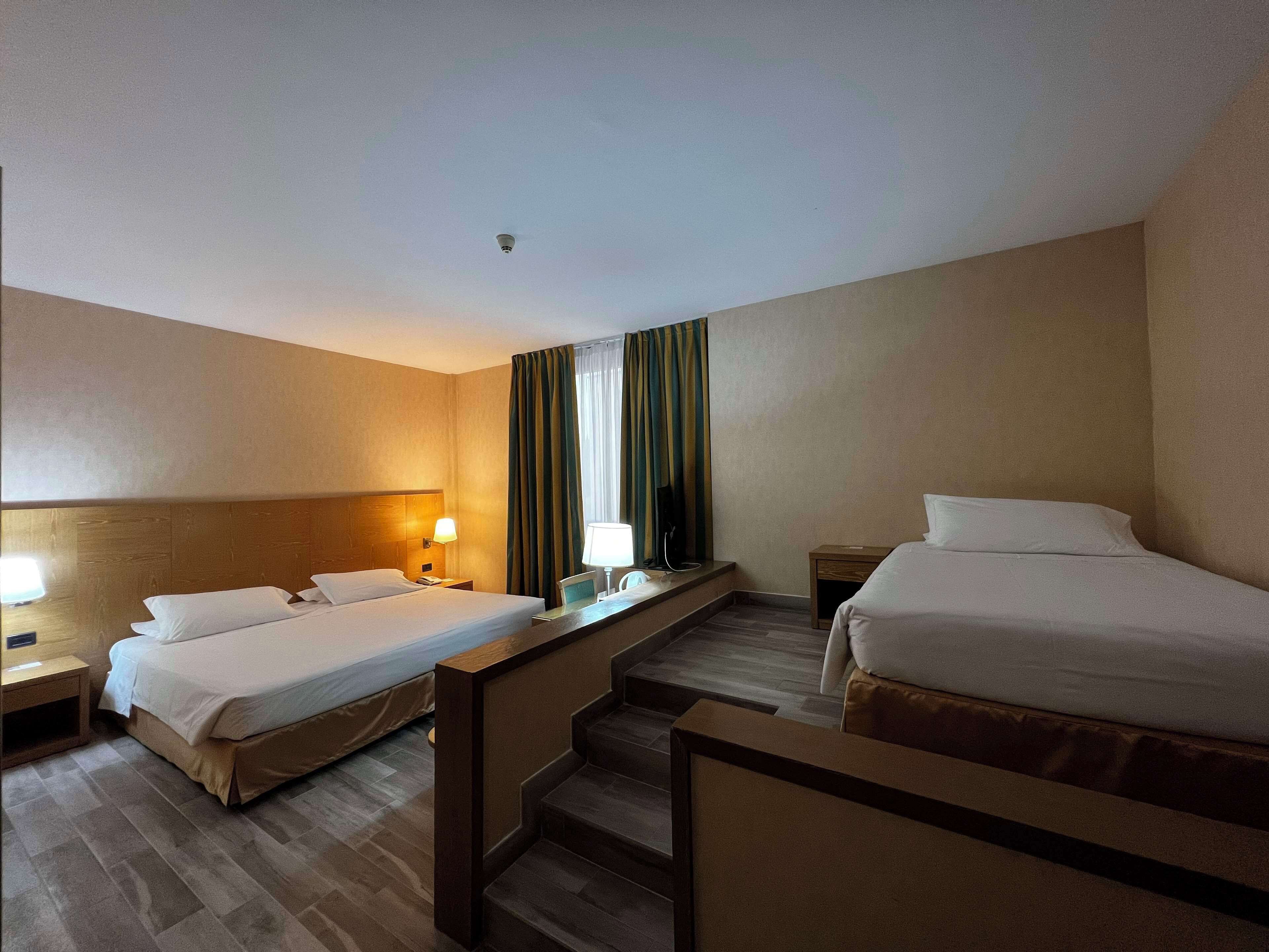 Quarto de hotel com duas camas, uma escrivaninha, uma cadeira, uma televisão e uma janela com cortinas.