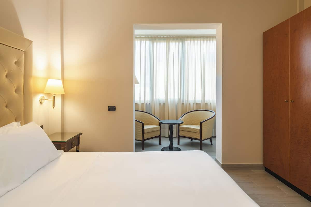 A hotel room with a bed, a nightstand, a lamp, a table with two chairs, and a wardrobe.