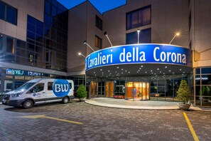 L'ingresso di un hotel con un cartello che recita 'Cavalieri della Corona', un furgone con la scritta 'BW Best Western' e una moderna facciata dell'edificio.
