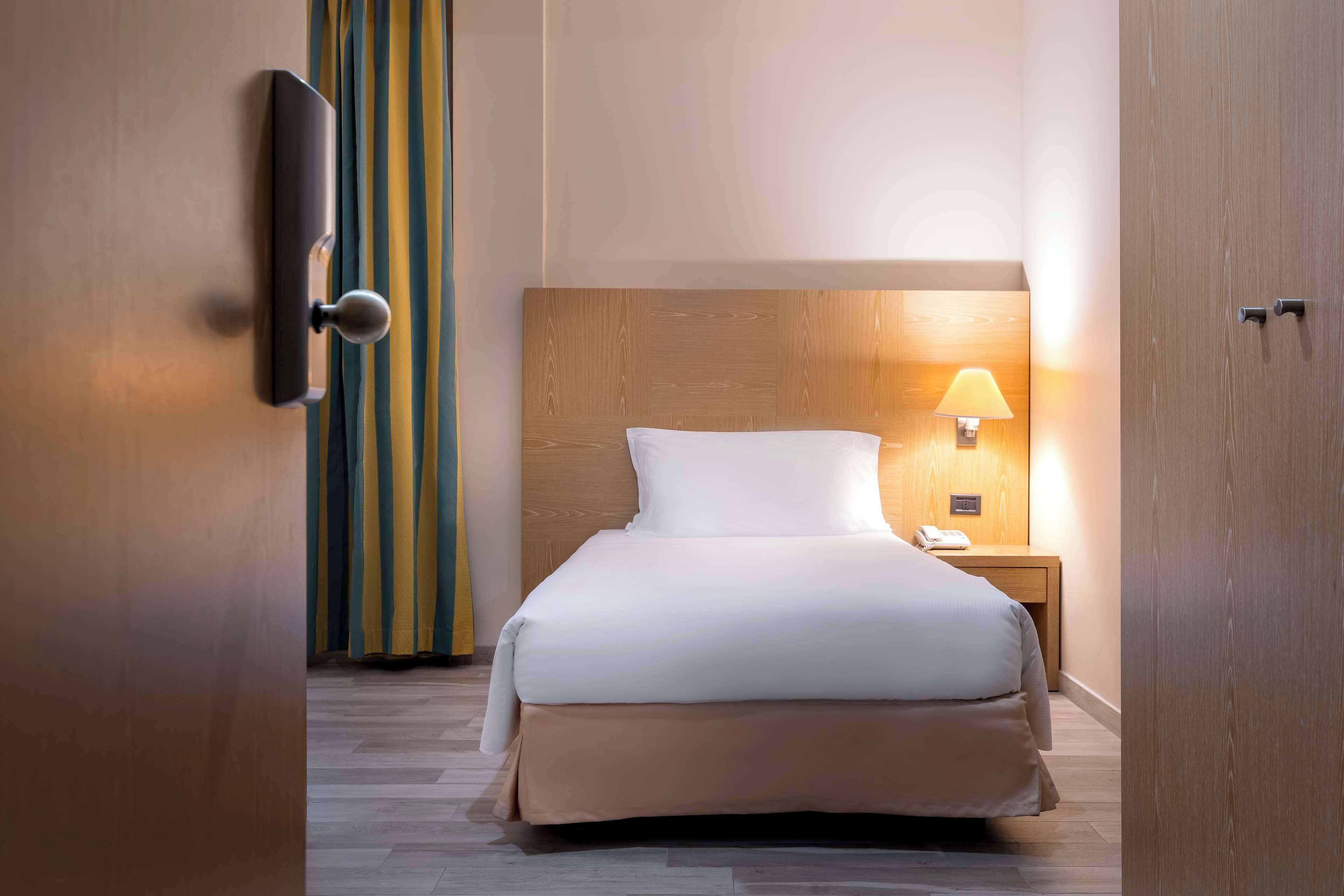 A hotel room with a wooden headboard, a bed with white bedding, a bedside lamp, and a wooden nightstand.