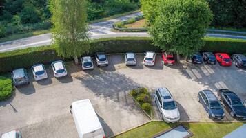 Vista aerea di un parcheggio con diverse auto e un furgone bianco, circondato da vegetazione e alberi.