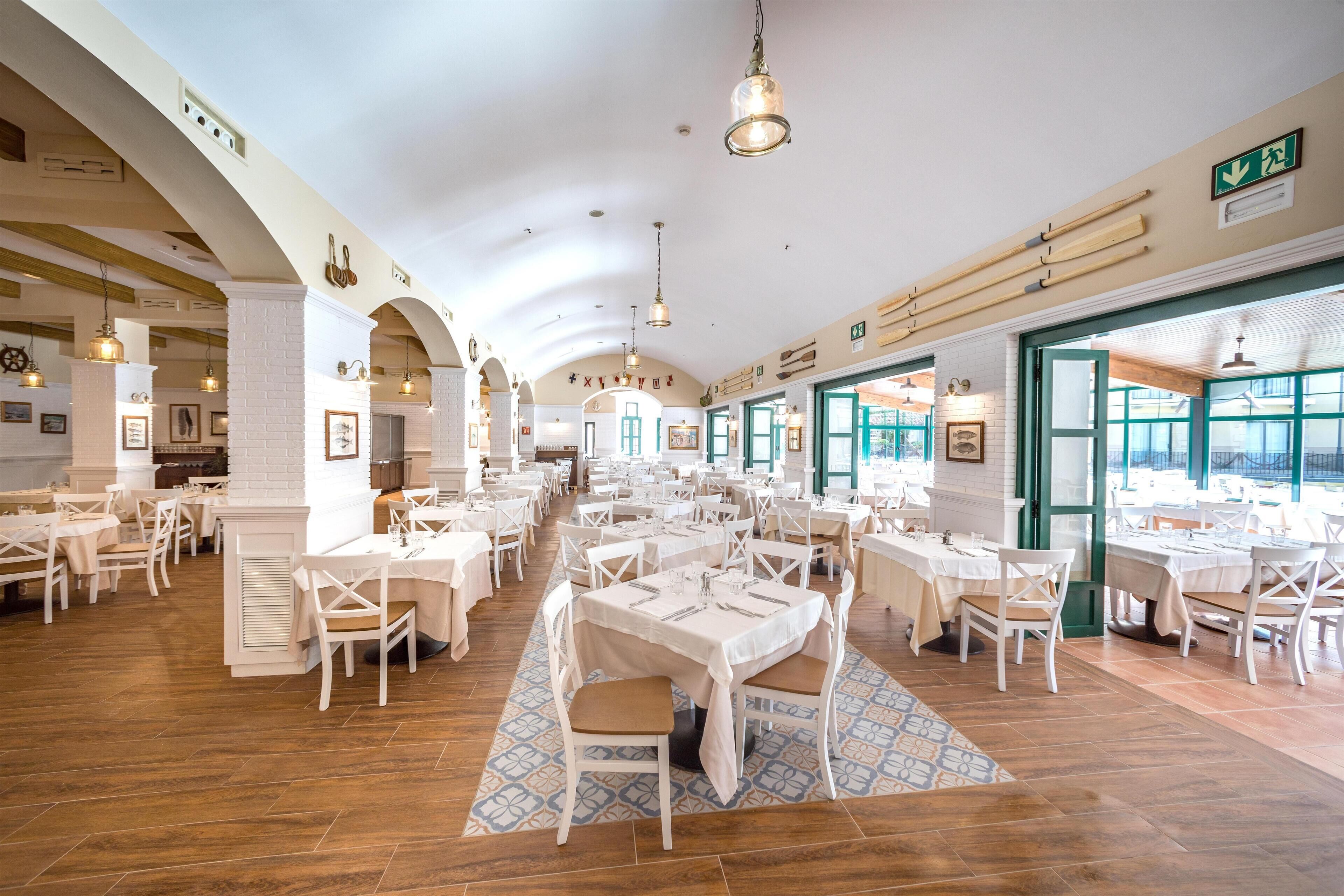 Un comedor espacioso con varias mesas y sillas, techos altos con vigas arqueadas y amplias ventanas que dejan entrar luz natural.